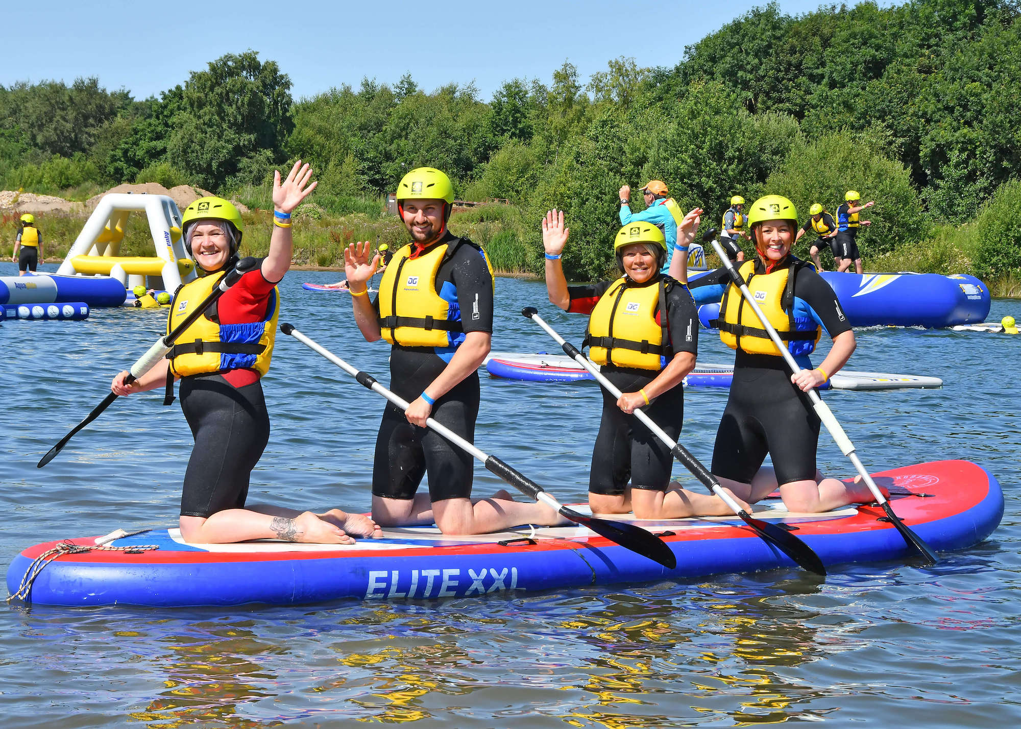 Four people knelt on one long paddle board in the water, waving at the camera