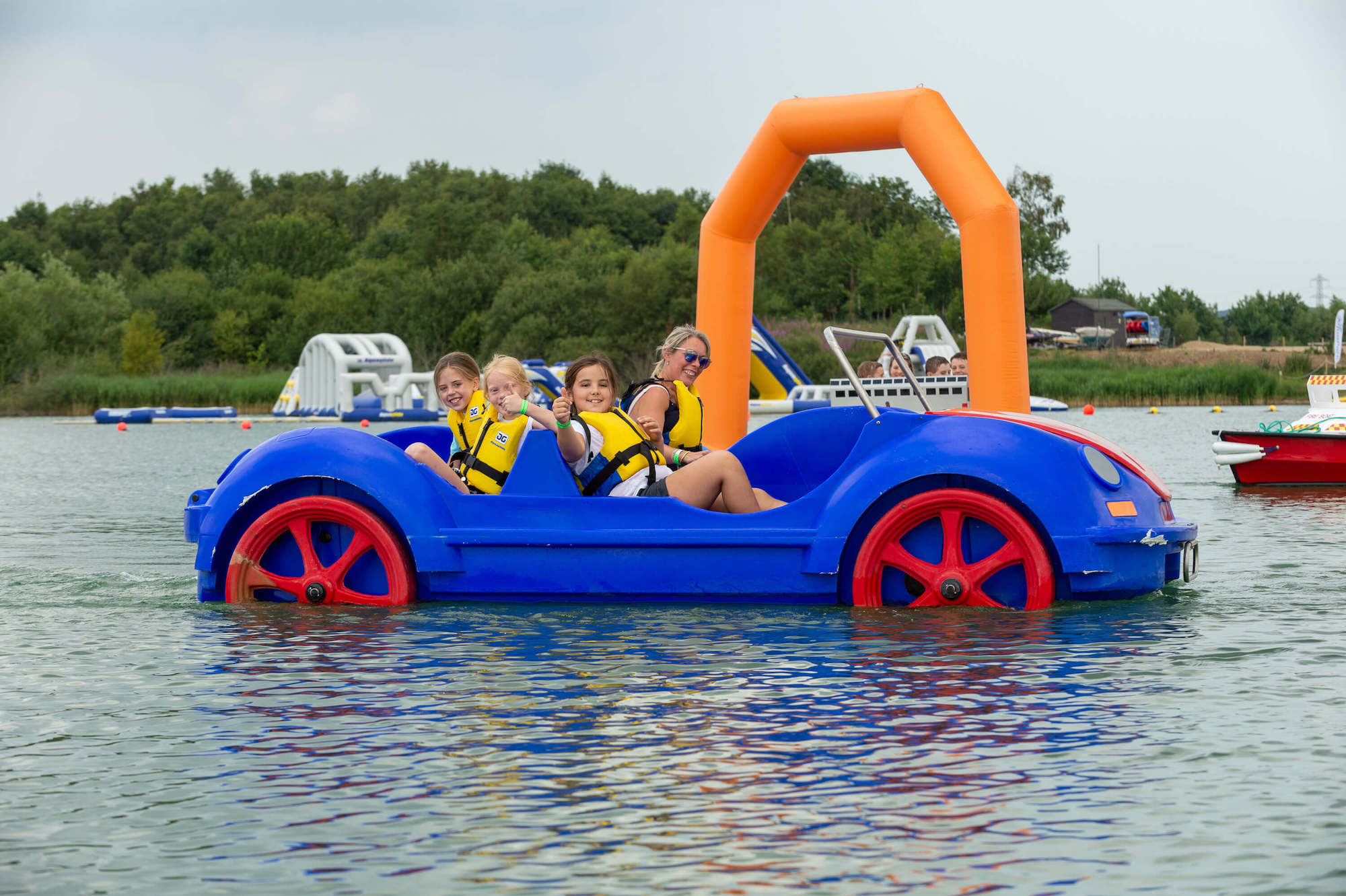 A large blue and red pedalo with a woman and three girls in it. One girl gives a thumbs up to the camera.