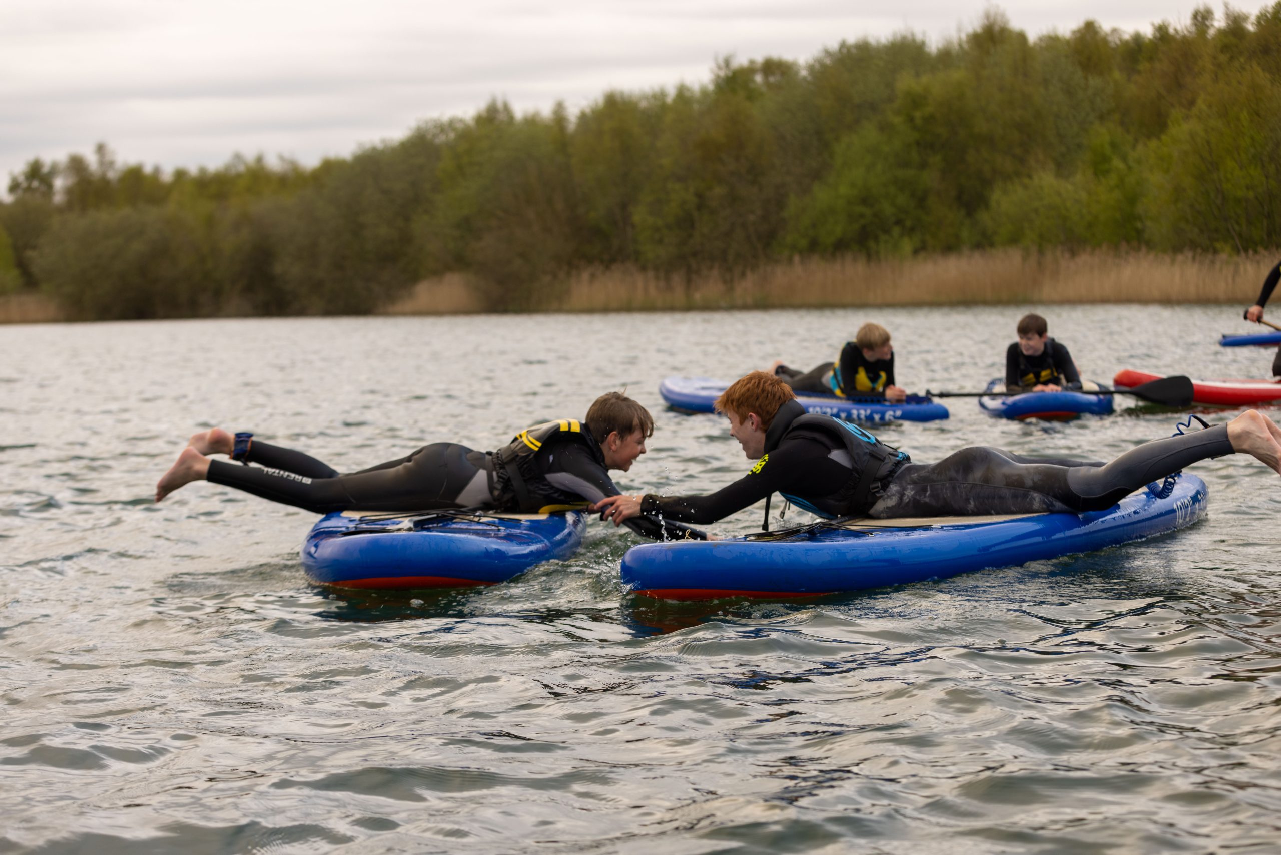 A group of children lay down on paddle boards in the middle of a lake surrounded by greenery and trees