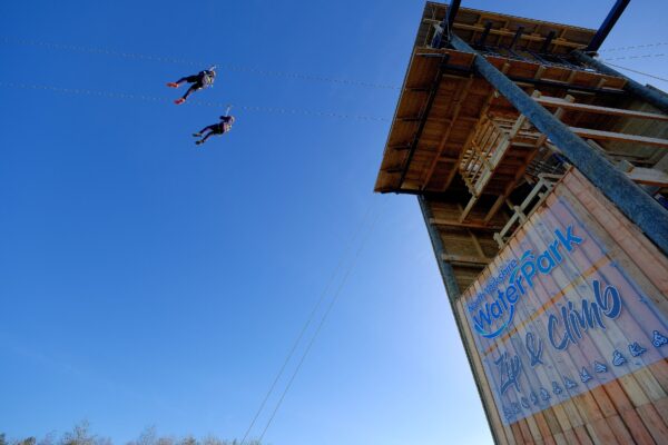 Two people in the air taking part in a zip line activity with a sign that says North Yorkshire Water Park Zip & Climb