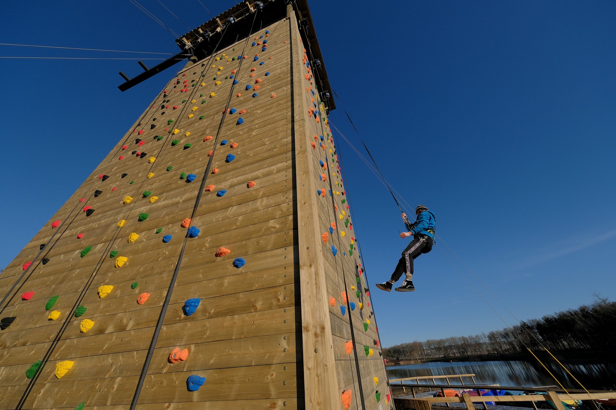 A person traverses a climbing wall at North Yorkshire Water Park with clear blue skies and a lake in the background