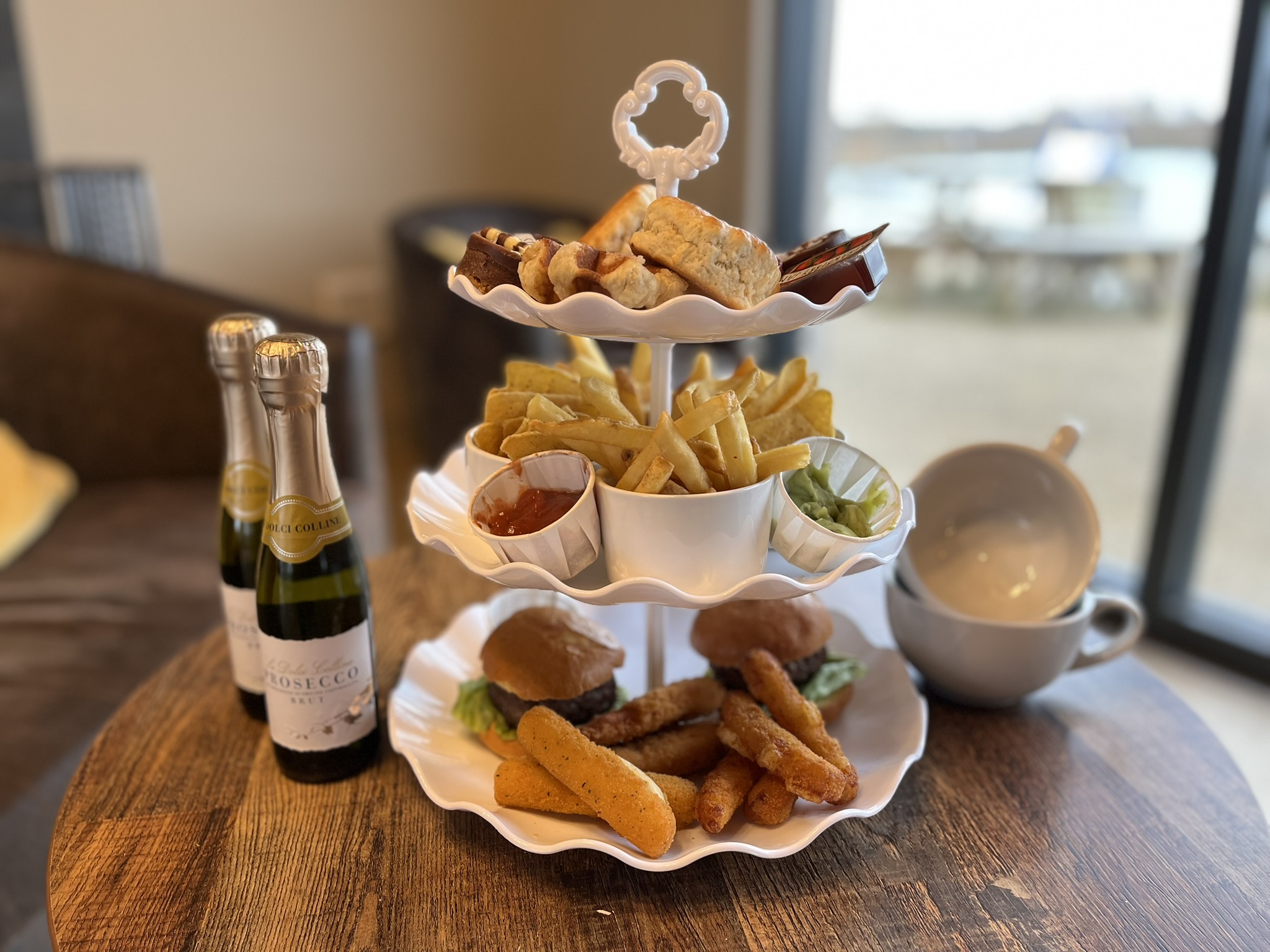 An afternoon tea stand with burgers, chips and sauces, along with two small bottles of champagne and coffee cups next to it
