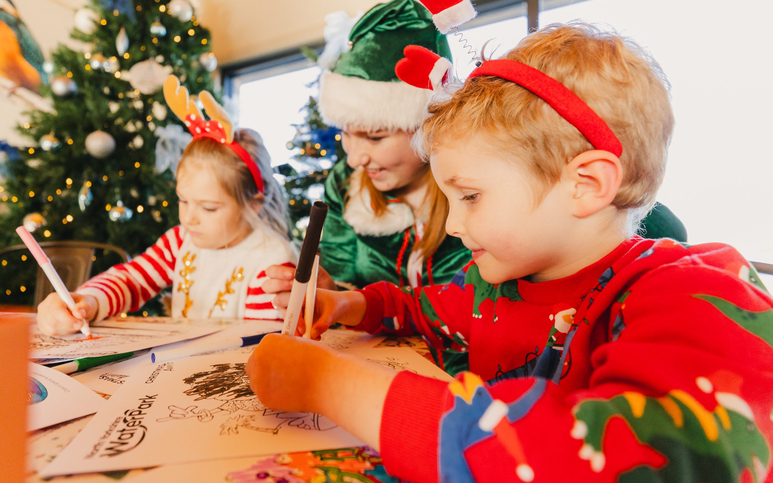Two children dressed in festive jumpers doing arts and crafts at a table with a woman dressed as an elf