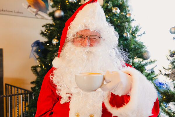 A man dressed as Santa Claus holds a coffee up in front of a Christmas tree covered in baubles