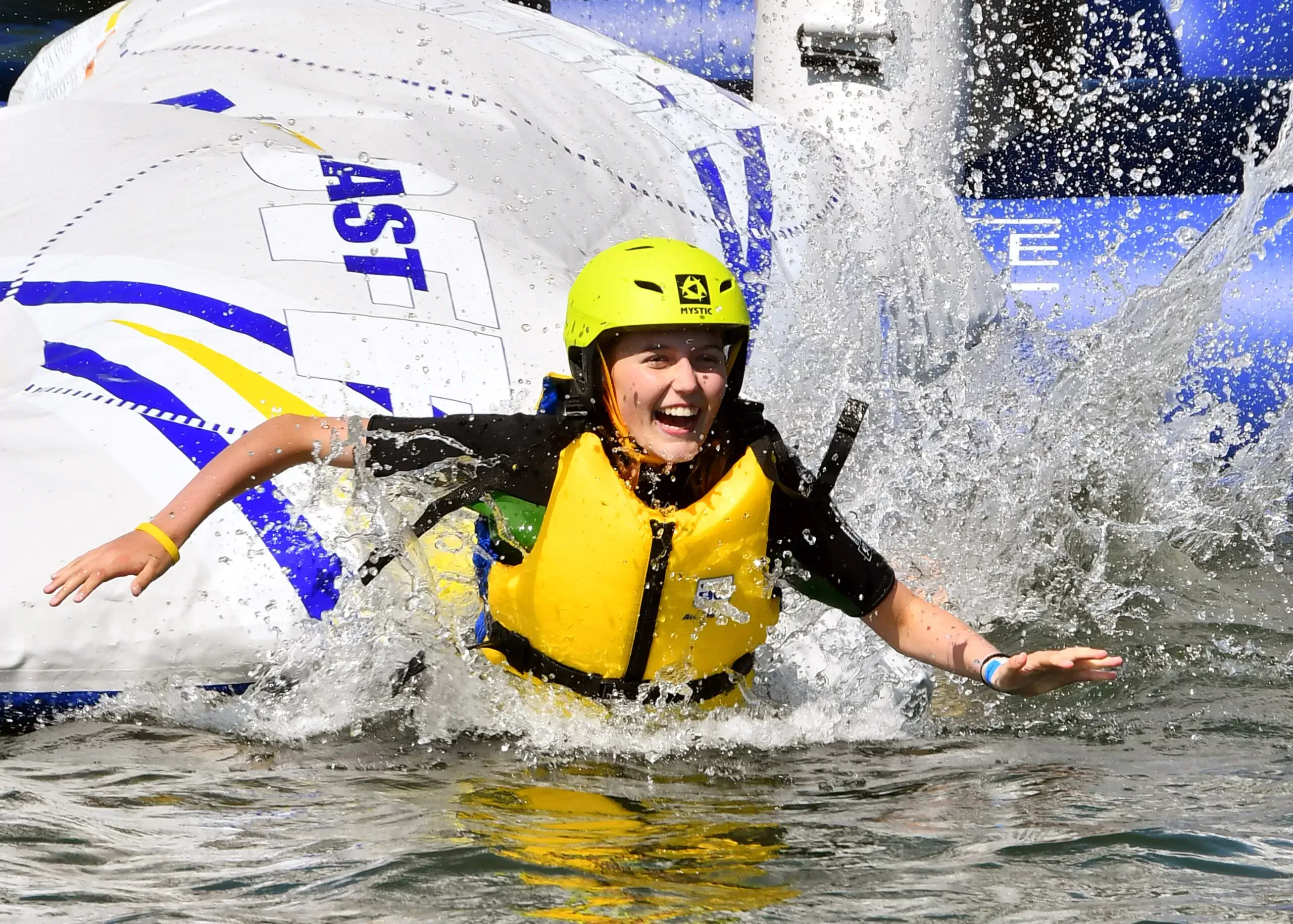 A grinning young woman splashes into a lake in a bright yellow helmet and vest after taking part in an inflatable AquaPark activity