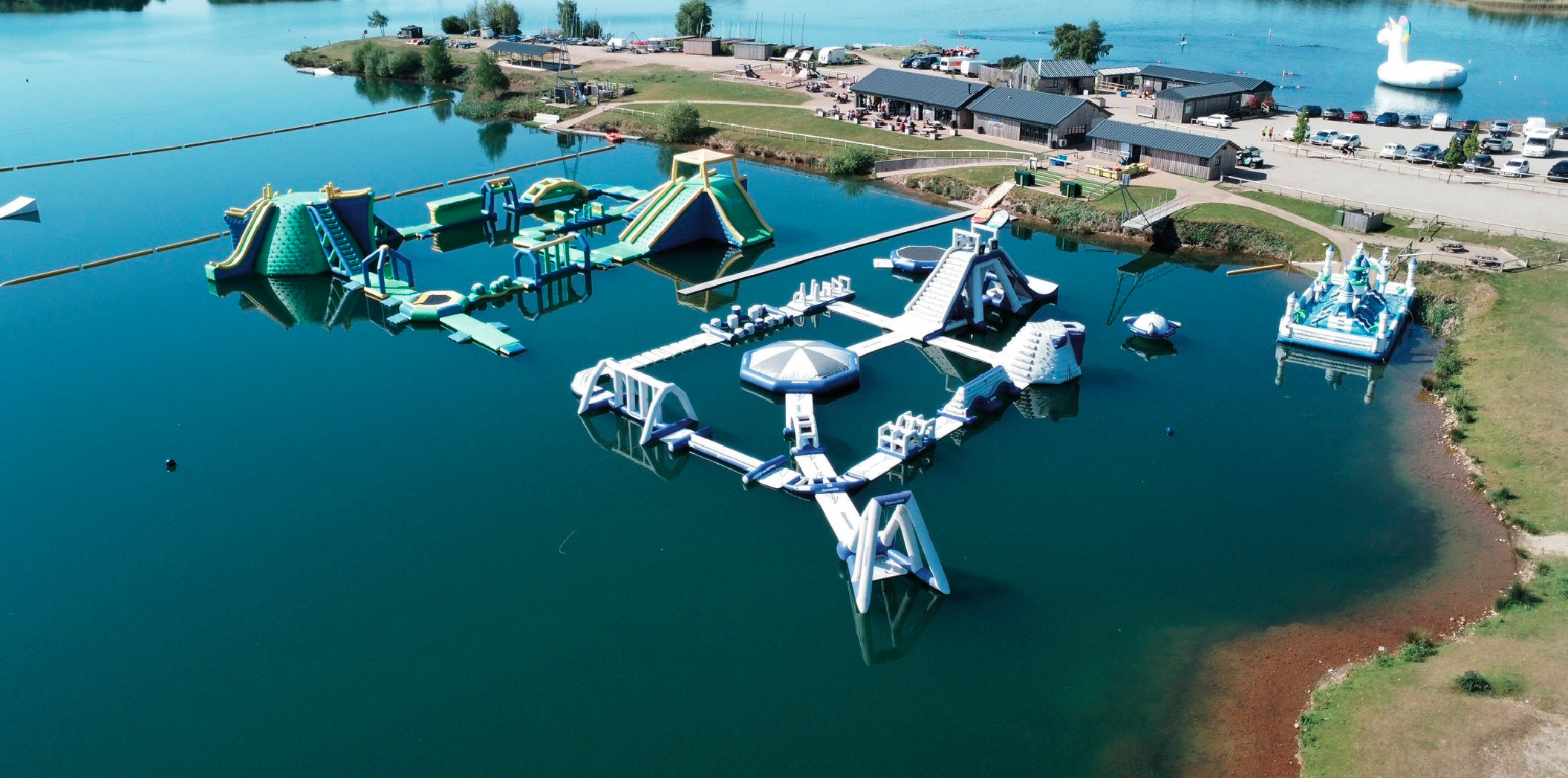 Wipeout and warrior inflatable aquaparks on a lake with cafe and reception buildings in background.
