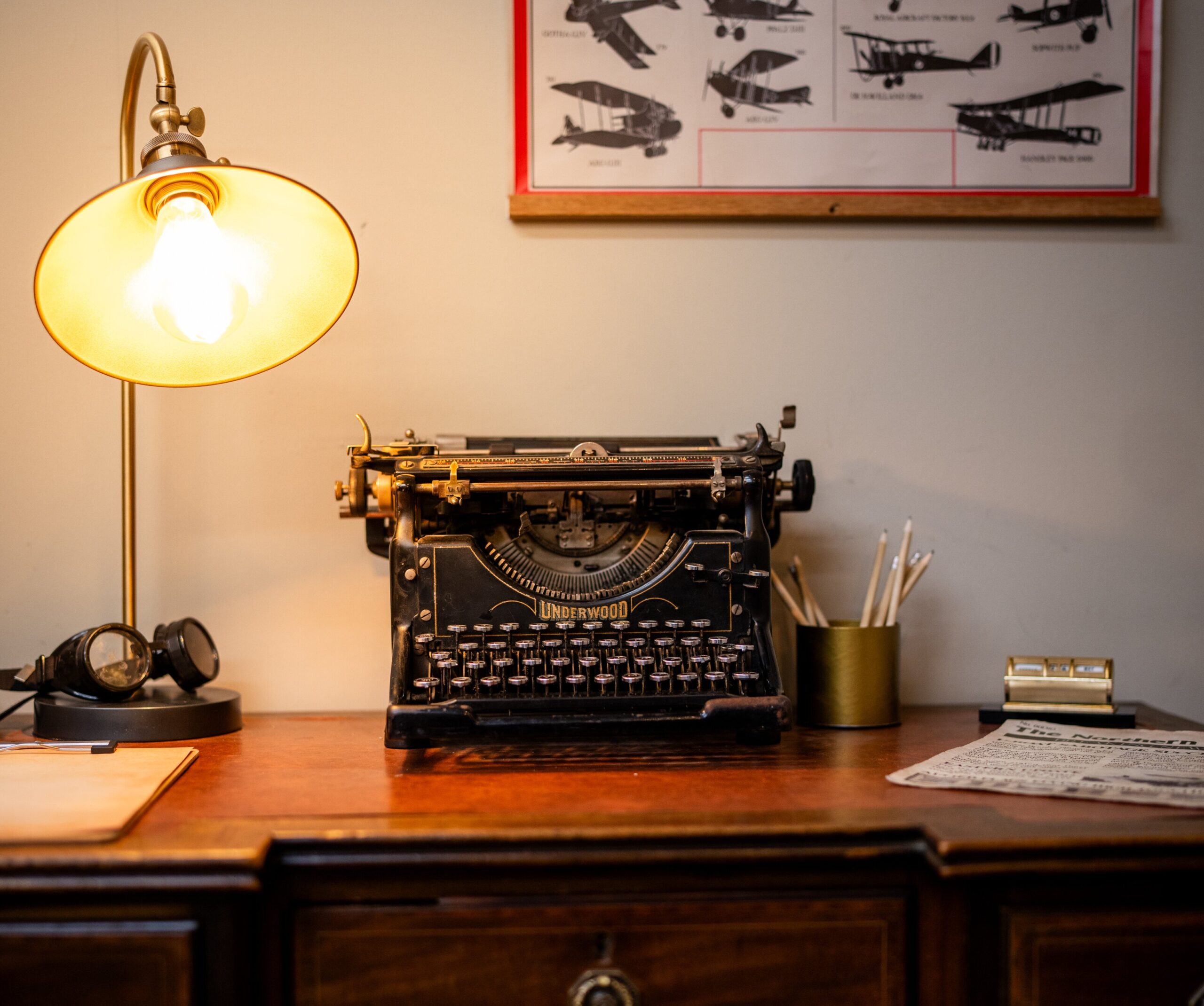 A typewriter on a wooden desk with a lamp shining above it and a picture of planes on the wall