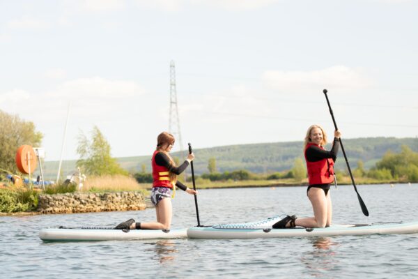Two people take part in stand up paddle boarding on a lake with greenery in the background
